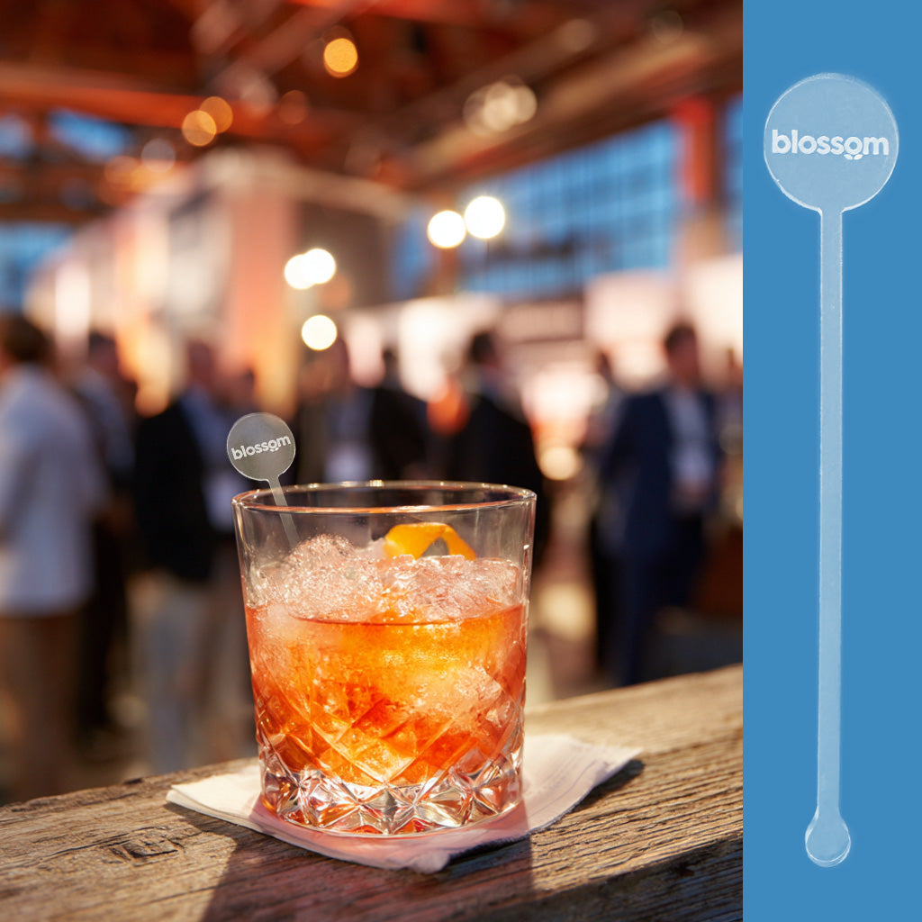 Cocktail in a glass with ice and an orange slice on a wooden bar, blurred background with Blossom branding. Blossom branded drink stirrer in a cocktail at a hotel bar corporate event