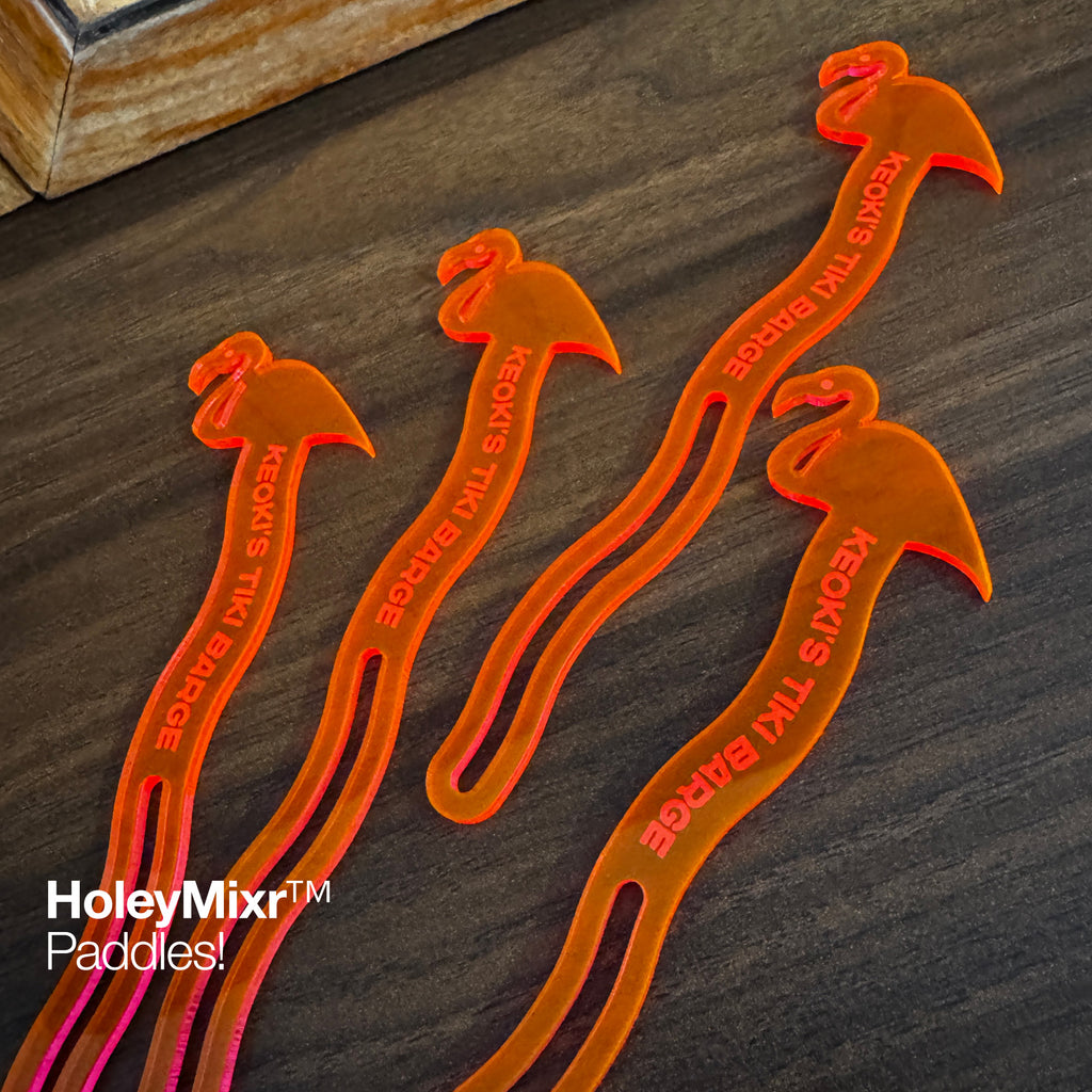 Flamingo Personalized Stubbie Swizzle Sticks with 'KEOKIS Tiki Barge' text, arranged on a dark wood surface with a logo overlay in the bottom left.