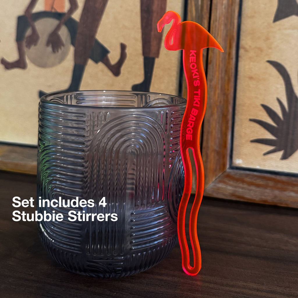 A textured dark gray glass tumbler on a wooden surface next to a Flamingo Personalized Stubbie Swizzle Stick with a flamingo-shaped handle and red text reading 'KEOKI'S TIKI STIRRER'. White text overlay says 'Set includes 4 Stubbie Stirrers'.