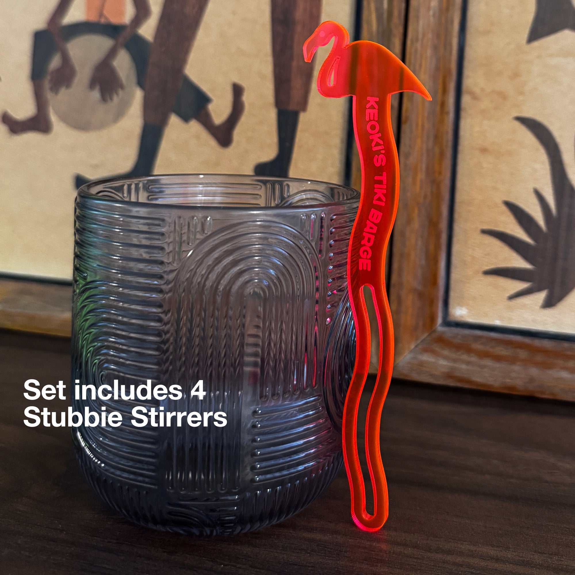 A textured dark gray glass tumbler on a wooden surface next to a Flamingo Personalized Stubbie Swizzle Stick with a flamingo-shaped handle and red text reading 'KEOKI'S TIKI STIRRER'. White text overlay says 'Set includes 4 Stubbie Stirrers'.