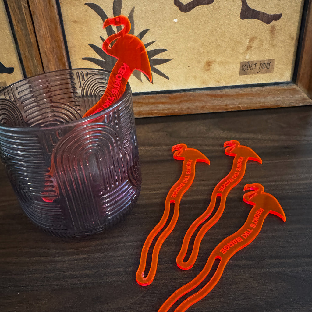 Flamingo Personalized Stubbie Swizzle Sticks designs are displayed on a dark wooden table next to a textured purple glass. The stirrers feature white text reading 'Keoki's Tiki Barge'