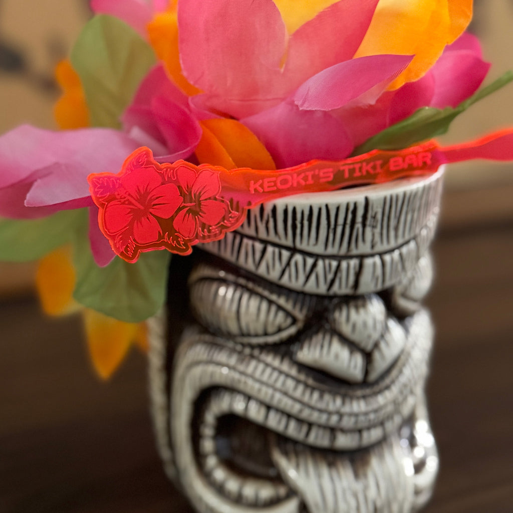 Tiki mug with colorful flowers and a Hibiscus Flower Personalized Stubbie Drink Stirrer with text on a blurred background