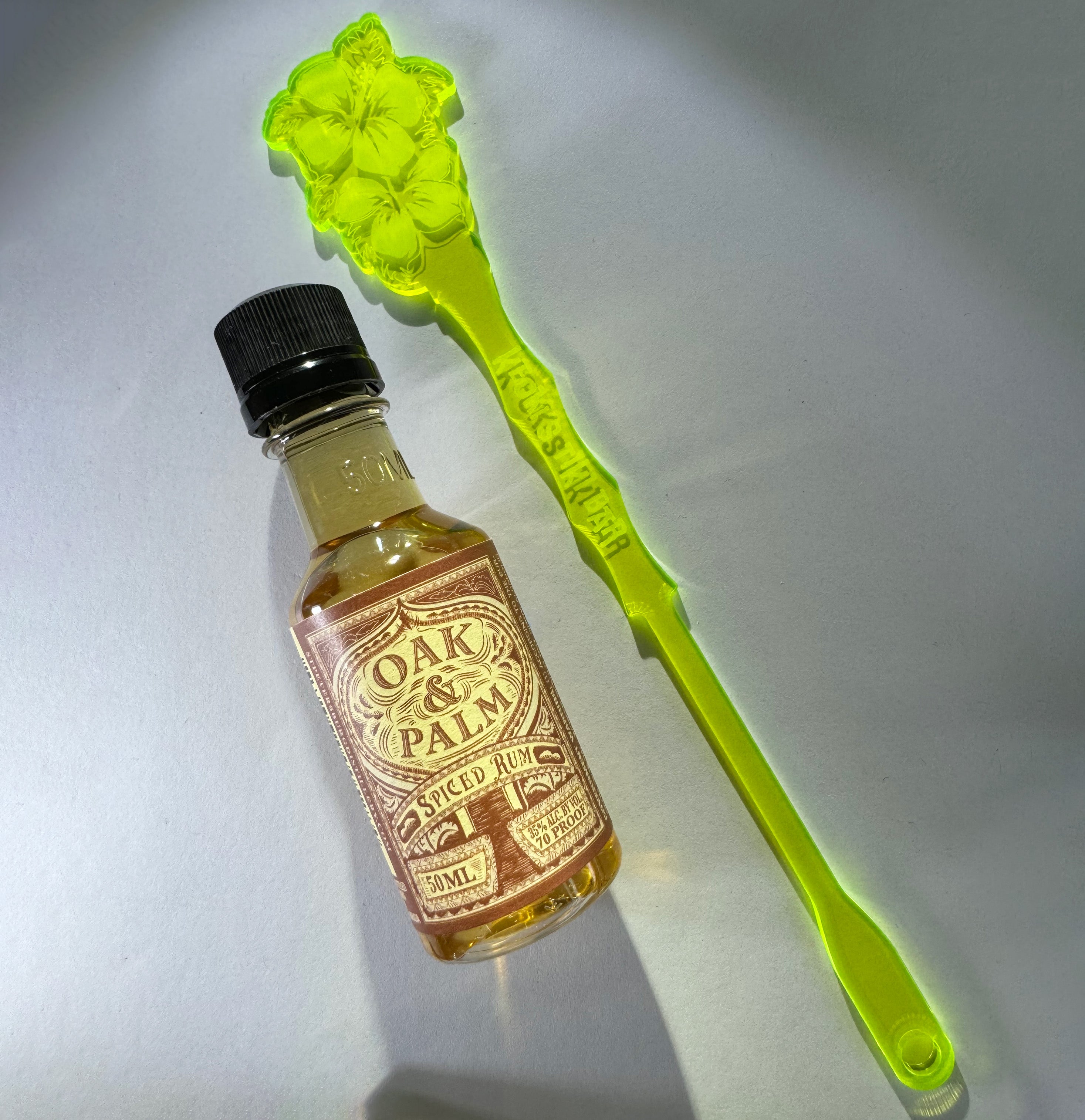 Small bottle of Oak & Palm Spiced Rum with a Hibiscus Flower Personalized XL Drink Stirrer on a light gray background