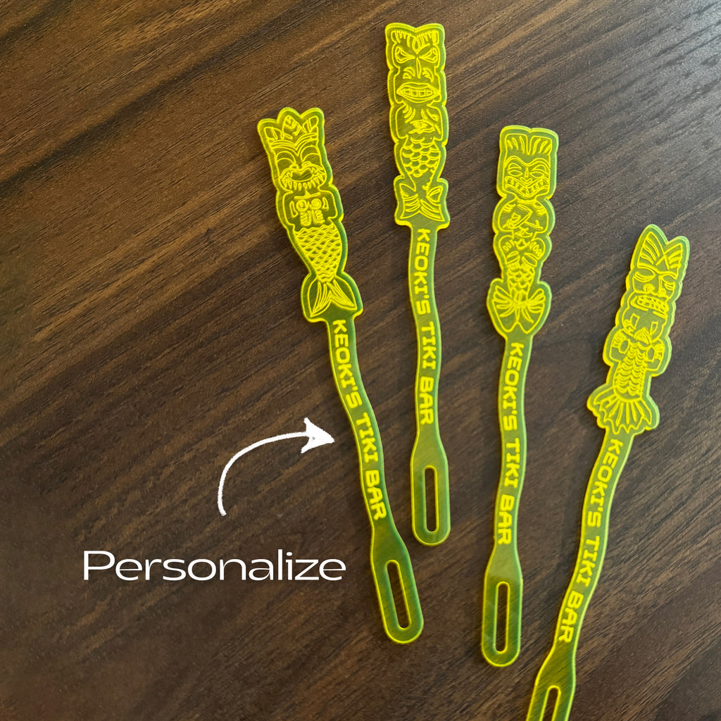 Mean Mermaids Personalized Stubbie Swizzle Sticks, acrylic cocktail stirrers with tiki designs and the text 'KEOKI'S TIKI BAR' printed on them, arranged on a dark wood surface. A white arrow points to one straw with the word 'Personalize' written below.