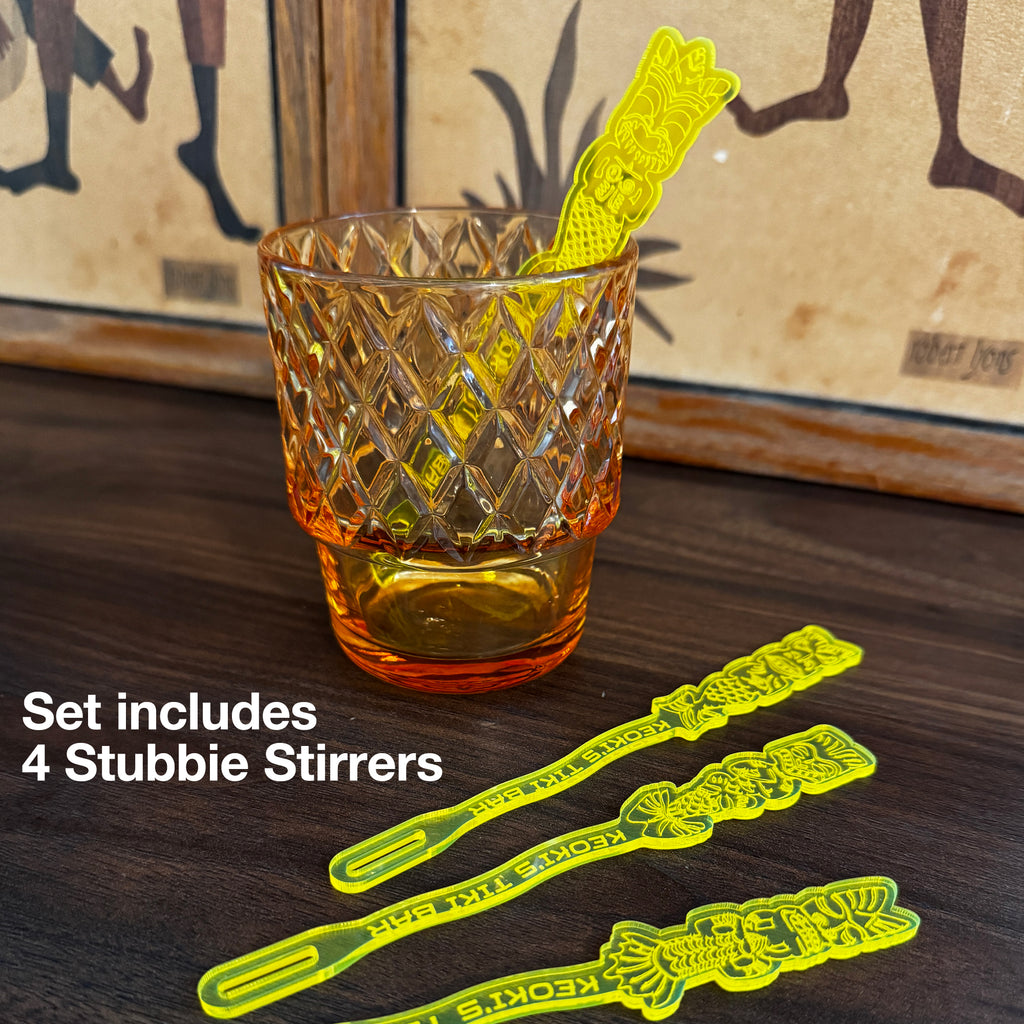 A clear ribbed tumbler glass with a Mean Mermaids Personalized Stubbie Swizzle Stick featuring a mean mermaid top and the text 'Keoki's Tiki Bar' printed vertically. White text on the image reads 'Set includes 4 Stubbie Stirrers.