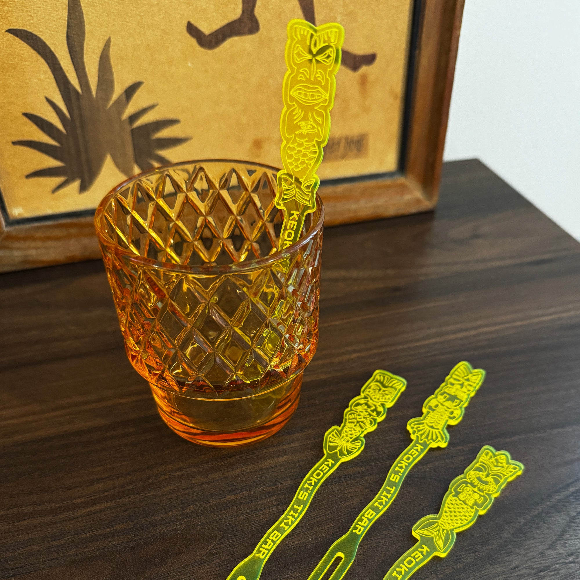 Mean Mermaids Personalized Stubbie Swizzle Sticks, tiki-style cocktail stirrers with carved mermaids and the words 'Keoki's Tiki Bar' resting on a dark wooden table next to an amber glass tumbler.