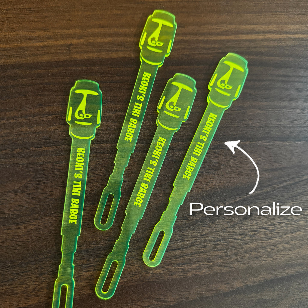 Moai Statue Stubbie Drink Stirrers with tiki designs and the text 'KEOKI'S TIKI BAR' printed on them, arranged on a dark wood surface. A white arrow points to one stirrer with the word 'Personalize' written below.