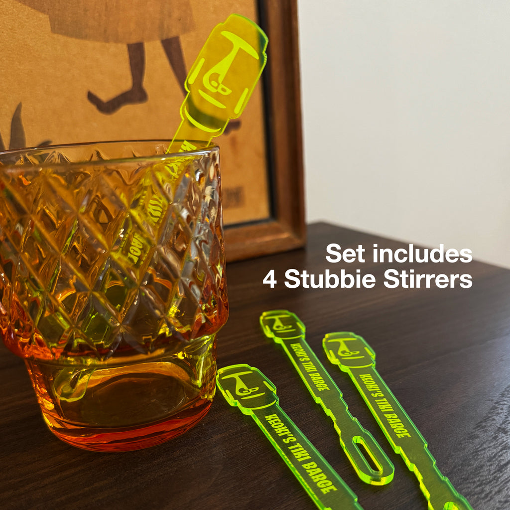 A textured glass tumbler on a wooden surface next to a Moai Statue Stubbie Drink Stirrer. White text overlay says 'Set includes 4 Stubbie Stirrers'.