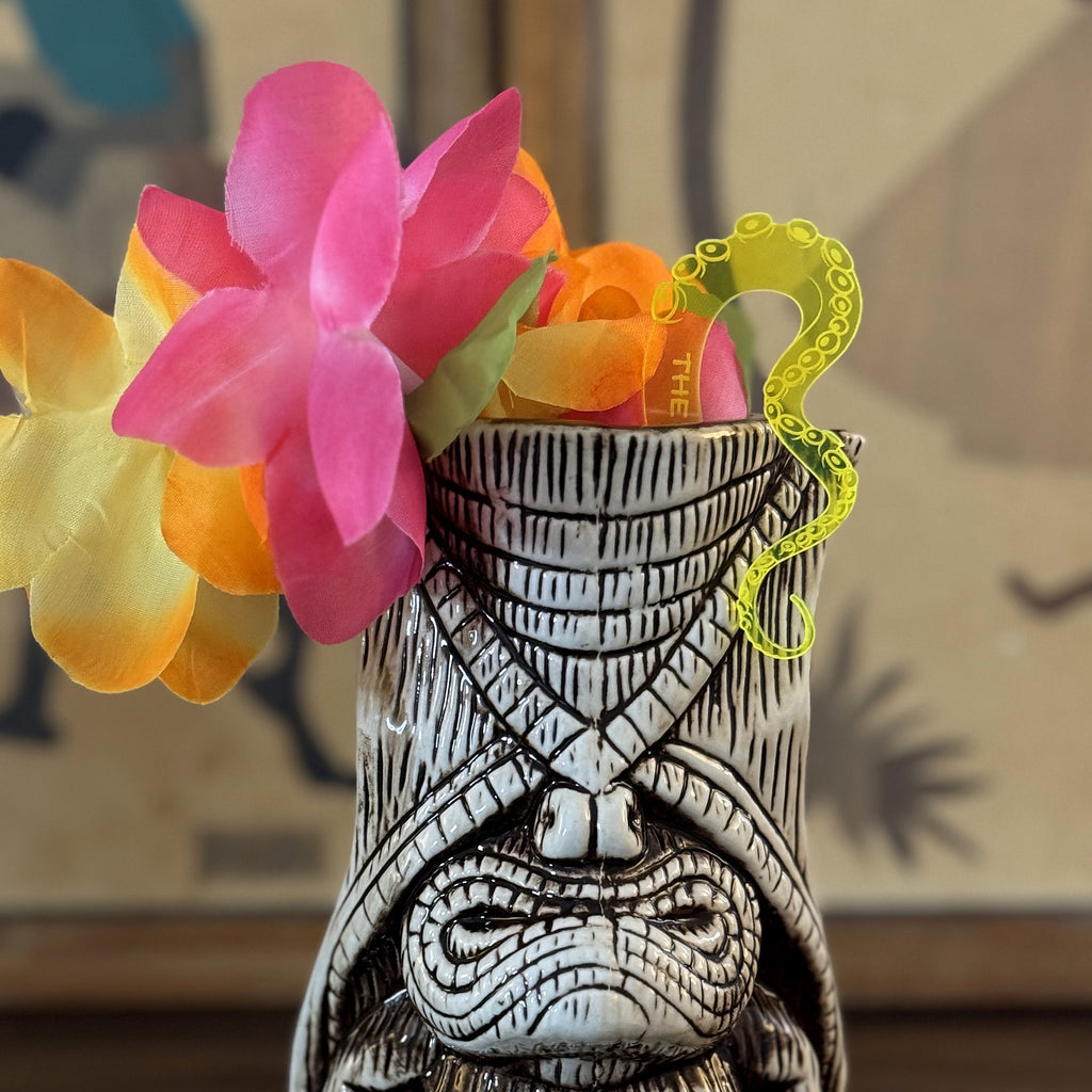 Octopus Tentacle "The Drunken Kraken" Personalized Stubbie Swizzle Stick in a Tiki mug and colorful flowers on a blurred background