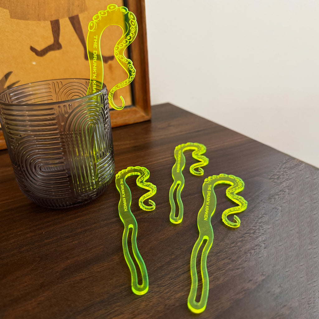 Octopus_Tentacle_The_Drunken_Kraken_Personalized_Stubbie_Swizzle_Sticks displayed on a dark wooden table next to a textured dark gray glass, with a framed print in the background.