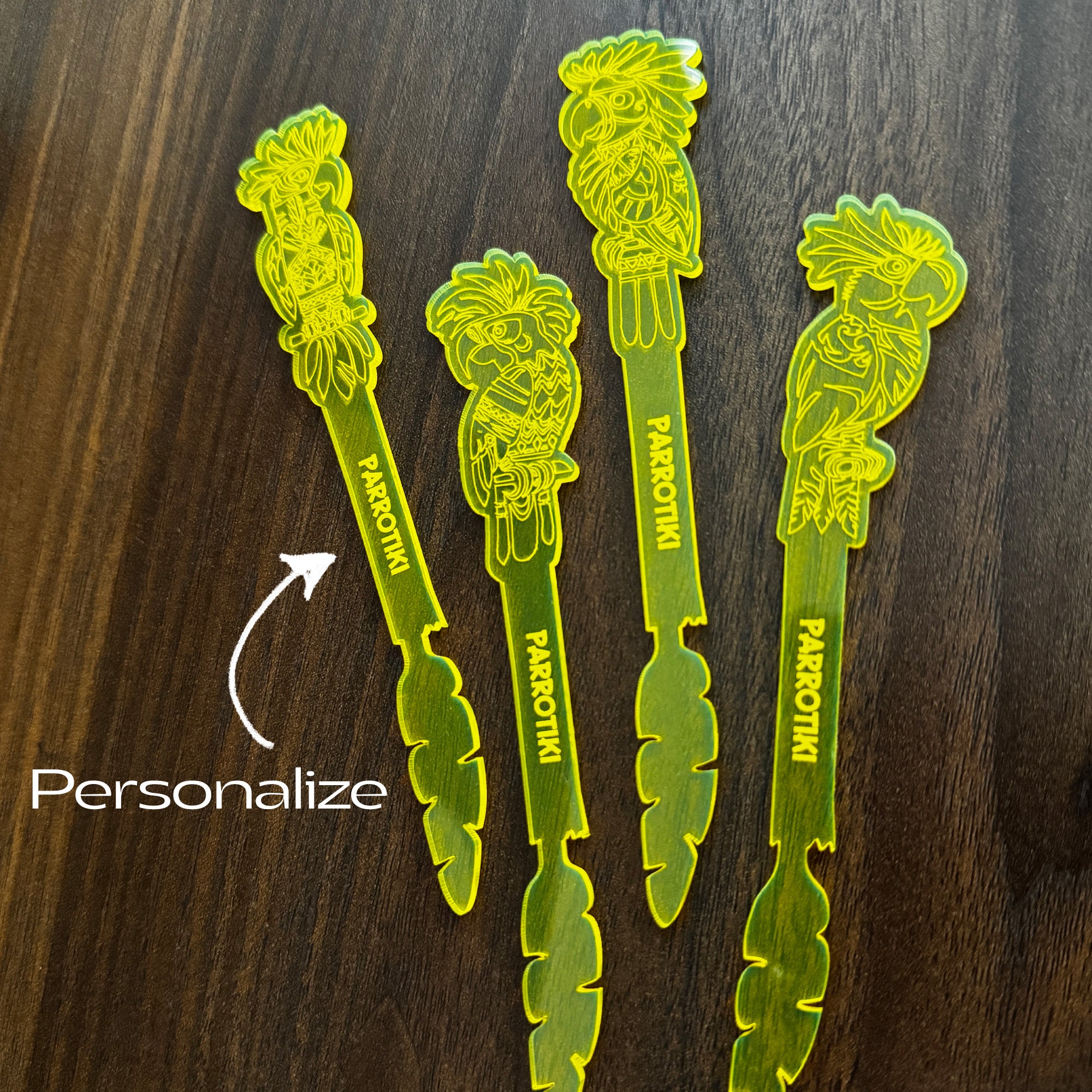 Parrotiki Cocktail Stubbie Stirrers with parrot designs and the name 'PARRPTIKII' printed vertically, arranged on a dark wood surface. A white arrow and the word 'Personalize' indicate customization.