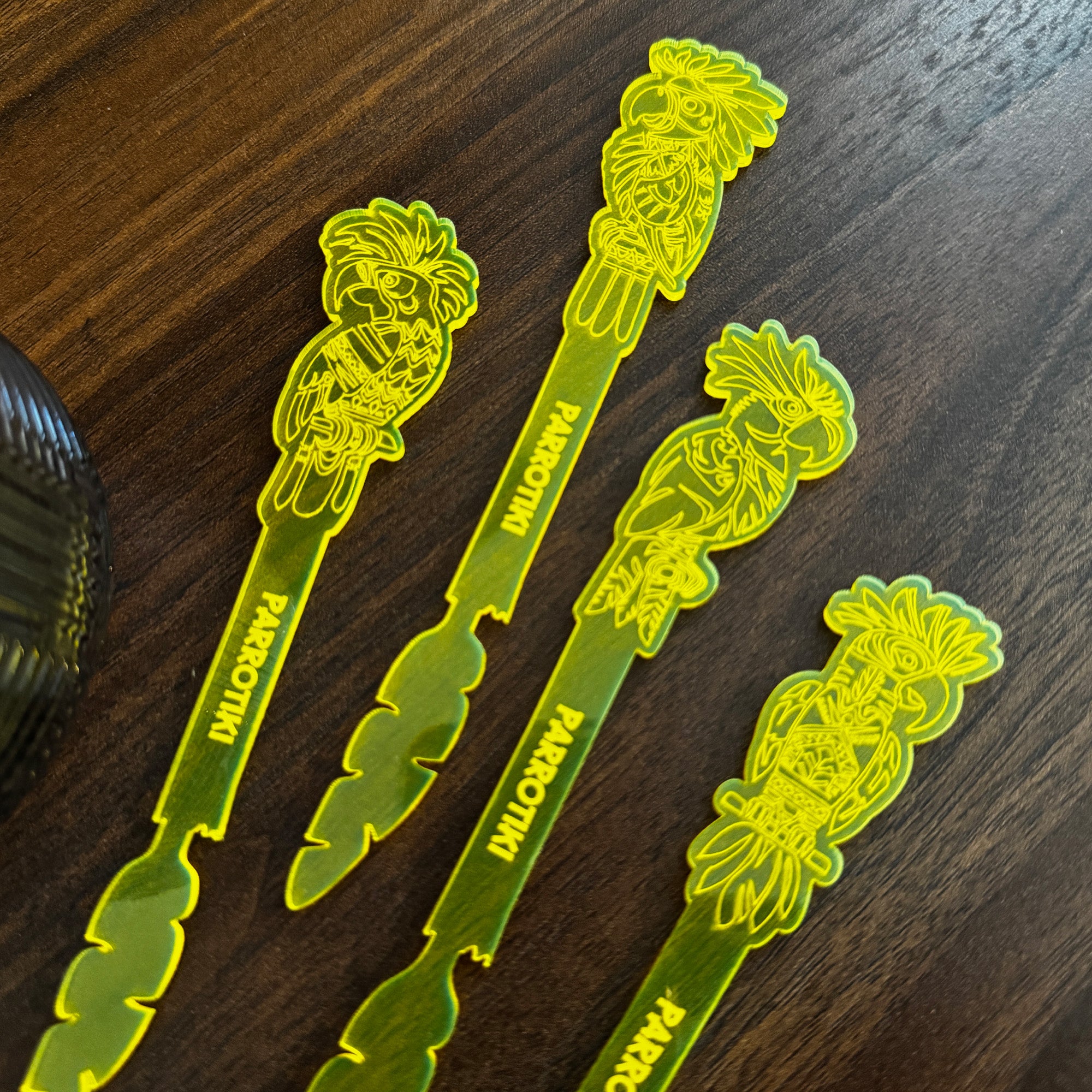 Parrotiki Cocktail Stubbie Stirrers, acrylic cocktail drink swizzle sticks featuring carved parrot designs and the word 'parrotiki' printed vertically, arranged on a dark wood tabletop.