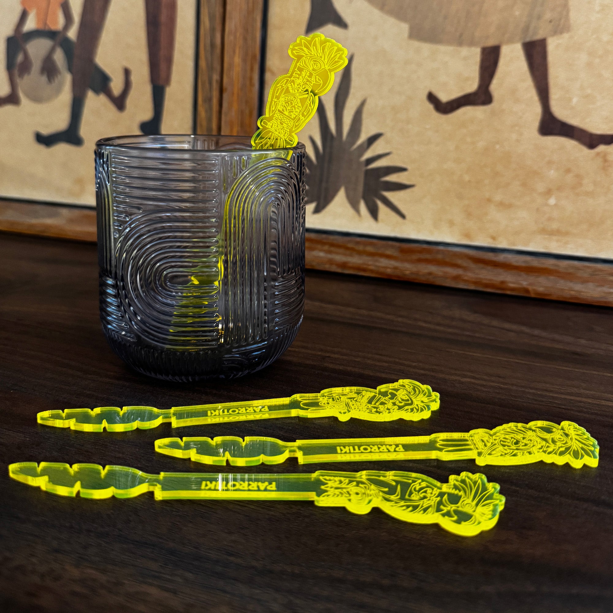 Parrotiki Cocktail Stubbie Stirrers are arranged on a dark wooden table, with a matching textured dark gray tumbler. The background features a warm-toned framed print with stylized figures.