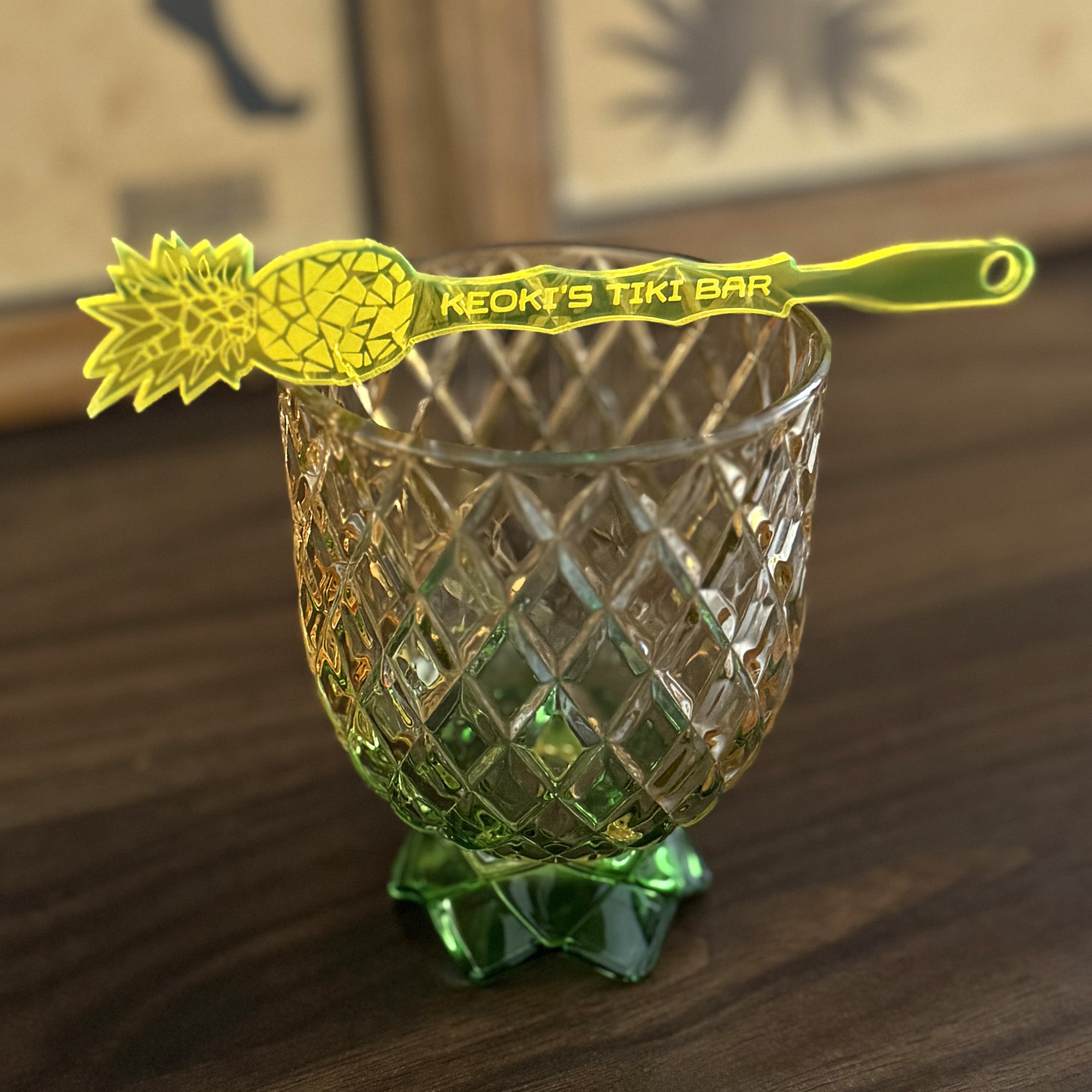 Decorative glass with pineapple design cocktail stirrer and 'Keoki's Tiki Bar' swizzle stick on a wooden surface