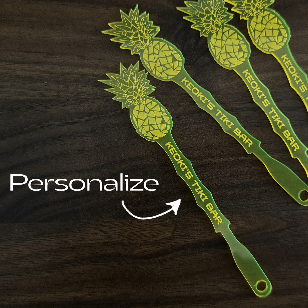 Pineapple shaped personalized drink stirrers with 'Keoki's Tiki Bar' text on a dark wooden surface, with the word 'Personalize' and an arrow pointing to one of the swizzle sticks.