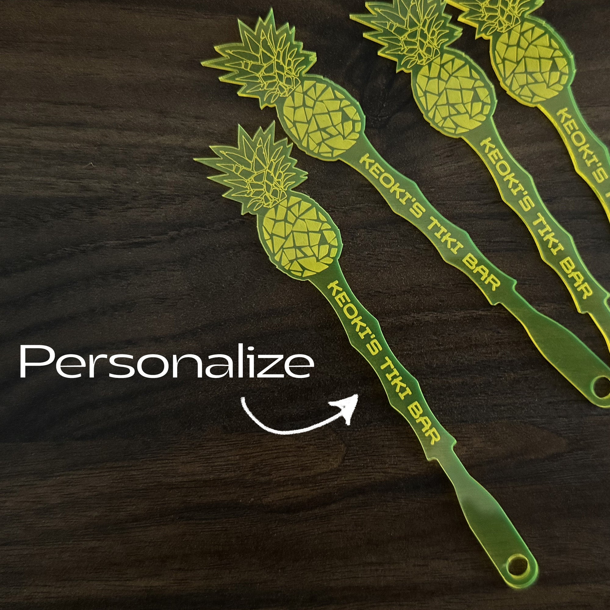 Pineapple shaped personalized drink stirrers with 'Keoki's Tiki Bar' text on a dark wooden surface, with the word 'Personalize' and an arrow pointing to one of the swizzle sticks.