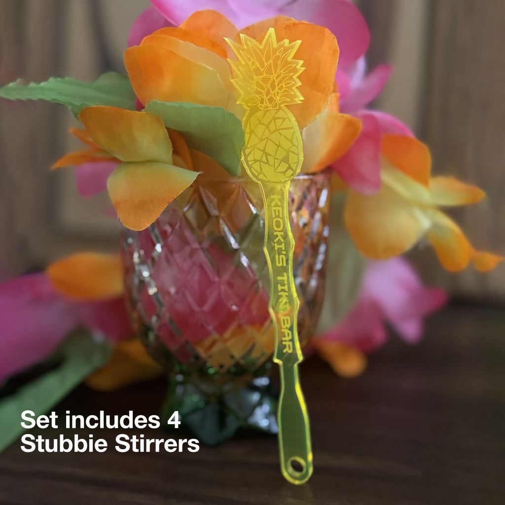 Pineapple shaped drink stirrers with a glass mug and flowers in the background.