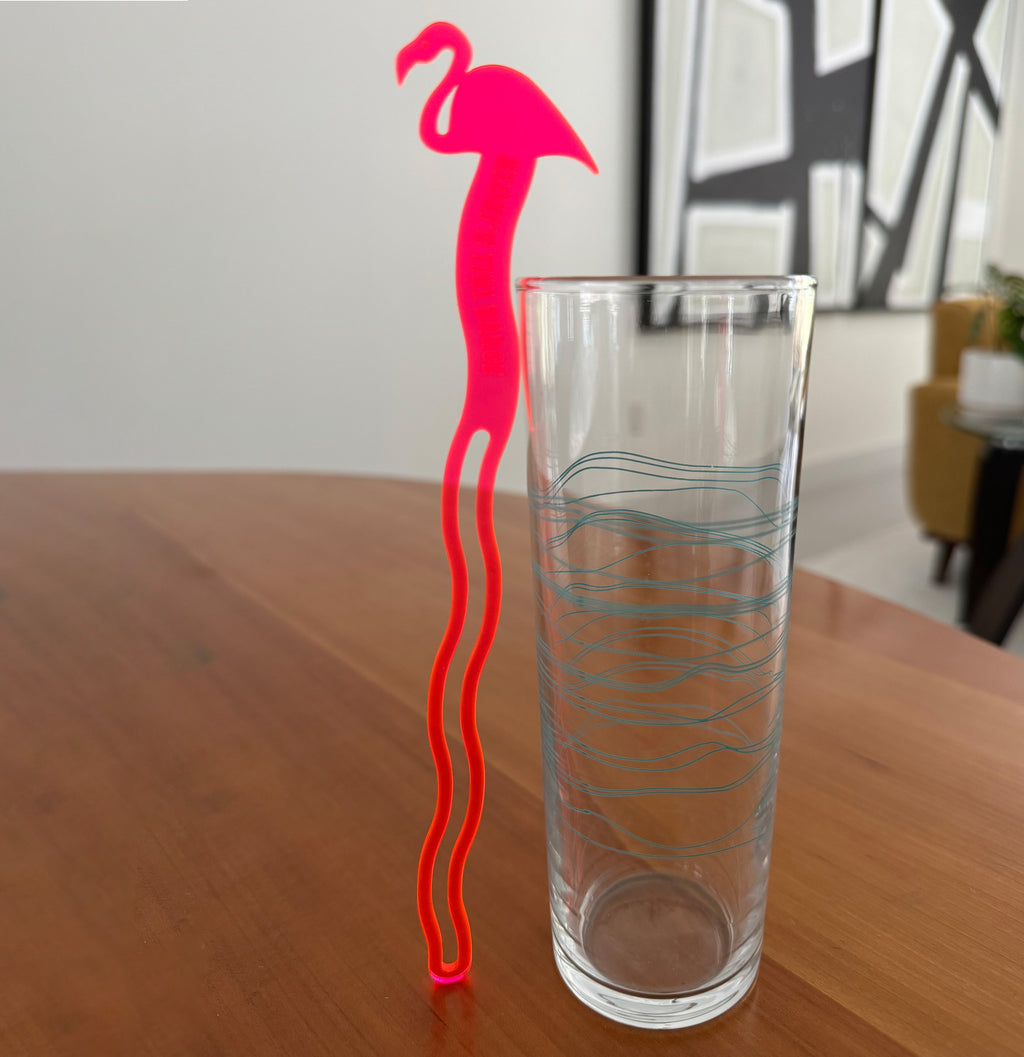 Clear glass with a Pink Flamingo Personalized Drink Stirrer on a wooden surface.