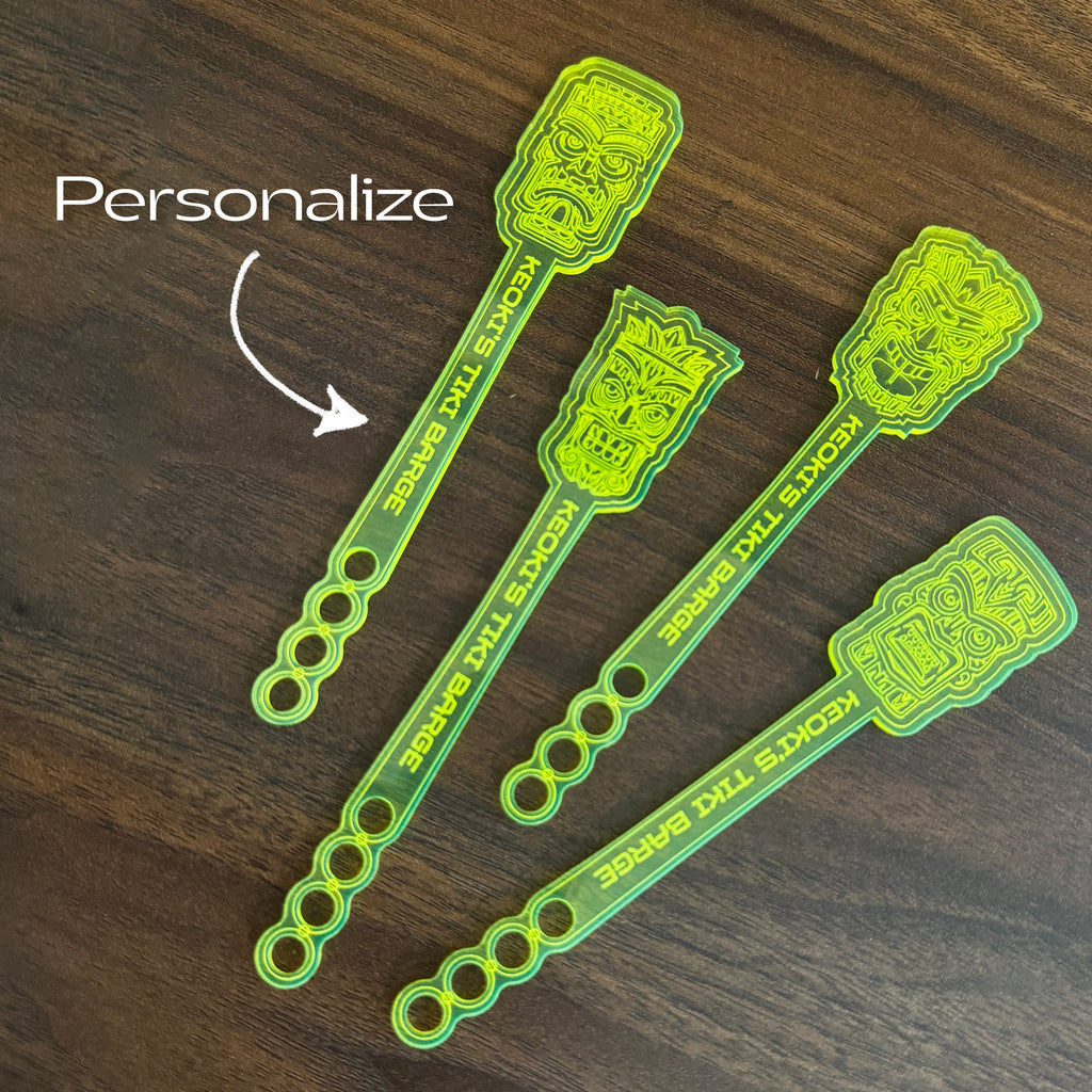 Four Tiki Neue Stubbie Drink Stirrers arranged on a dark wood background, with a white 'Personalize' text overlay and arrow pointing to one of the stirrers.