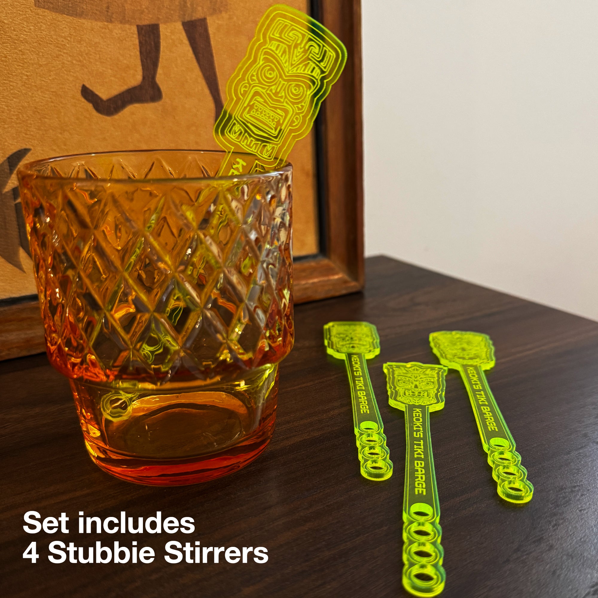 A textured amber glass tumbler on a wooden surface next to a Tiki Neue Stubbie Drink Stirrer . White text overlay says 'Set includes 4 Stubbie Stirrers'.