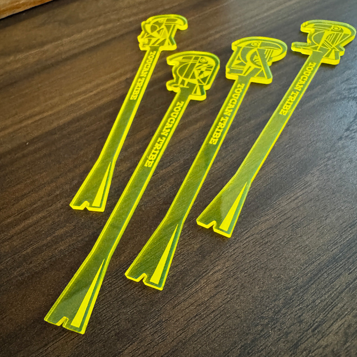 Four translucent neon yellow acrylic toucan logo cocktail drink stirrers with 'TOUCAN TRIBE' text and toucan logo art designs, arranged on a dark wood surface.