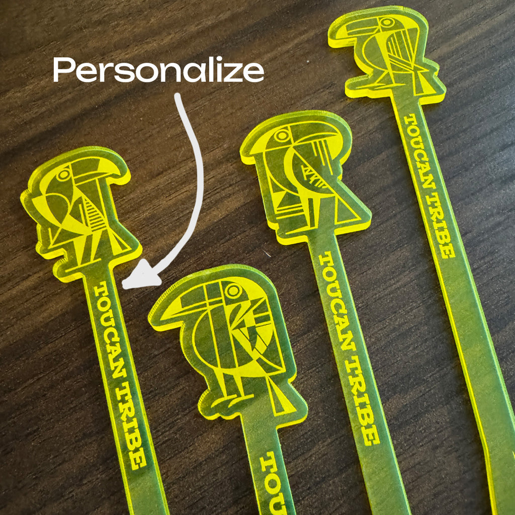 Four green and yellow Toucan Tribe acrylic logo cocktail drink stirrers on a dark wood background, with a white arrow and the word 'Personalize' indicating customization.