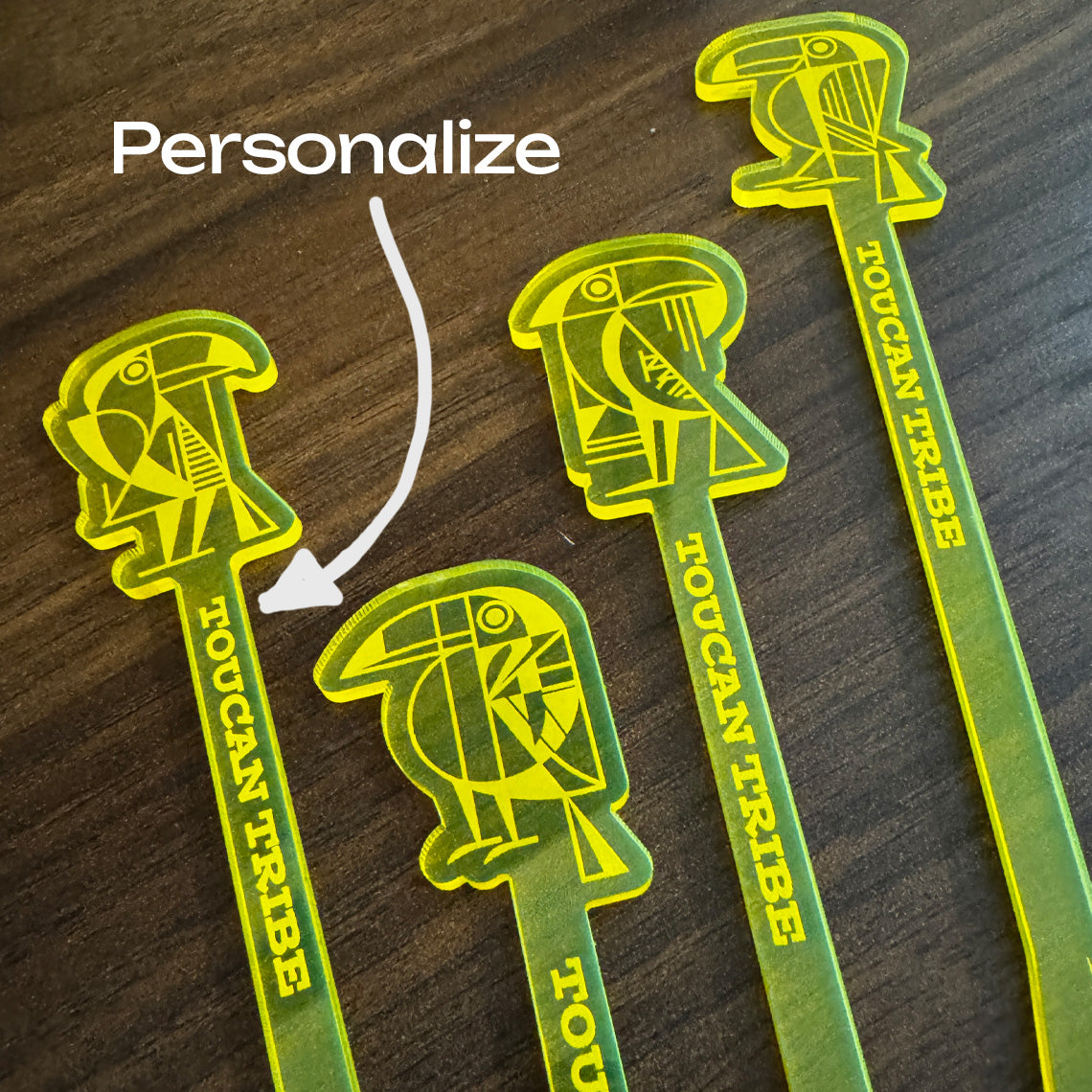 Four green and yellow Toucan Tribe acrylic logo cocktail drink stirrers on a dark wood background, with a white arrow and the word 'Personalize' indicating customization.