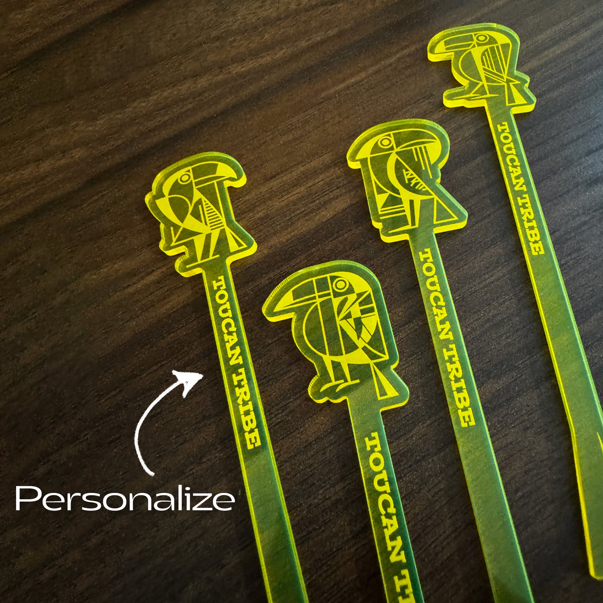 Four green and yellow Toucan Tribe acrylic logo cocktail drink stirrers on a dark wood background, with a white arrow and the word 'Personalize' indicating customization.