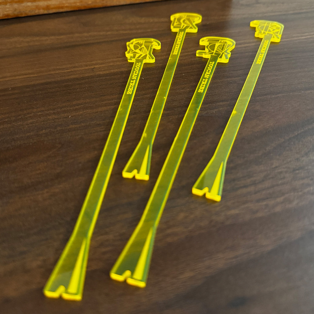 Four translucent yellow plastic cocktail straws with Toucan logo art designs on the tops, arranged on a dark wood surface.