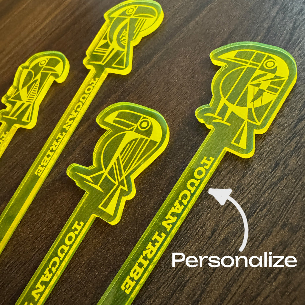 Four translucent neon yellow cocktail straws with Toucan Tribe logos and the words 'TOUCAN TRIBE' printed on them, arranged on a dark wood surface. A white arrow points to one straw with the word 'Personalize' overlaid.
