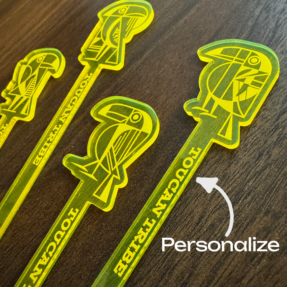 Four translucent neon yellow cocktail straws with Toucan Tribe logos and the words 'TOUCAN TRIBE' printed on them, arranged on a dark wood surface. A white arrow points to one straw with the word 'Personalize' overlaid.
