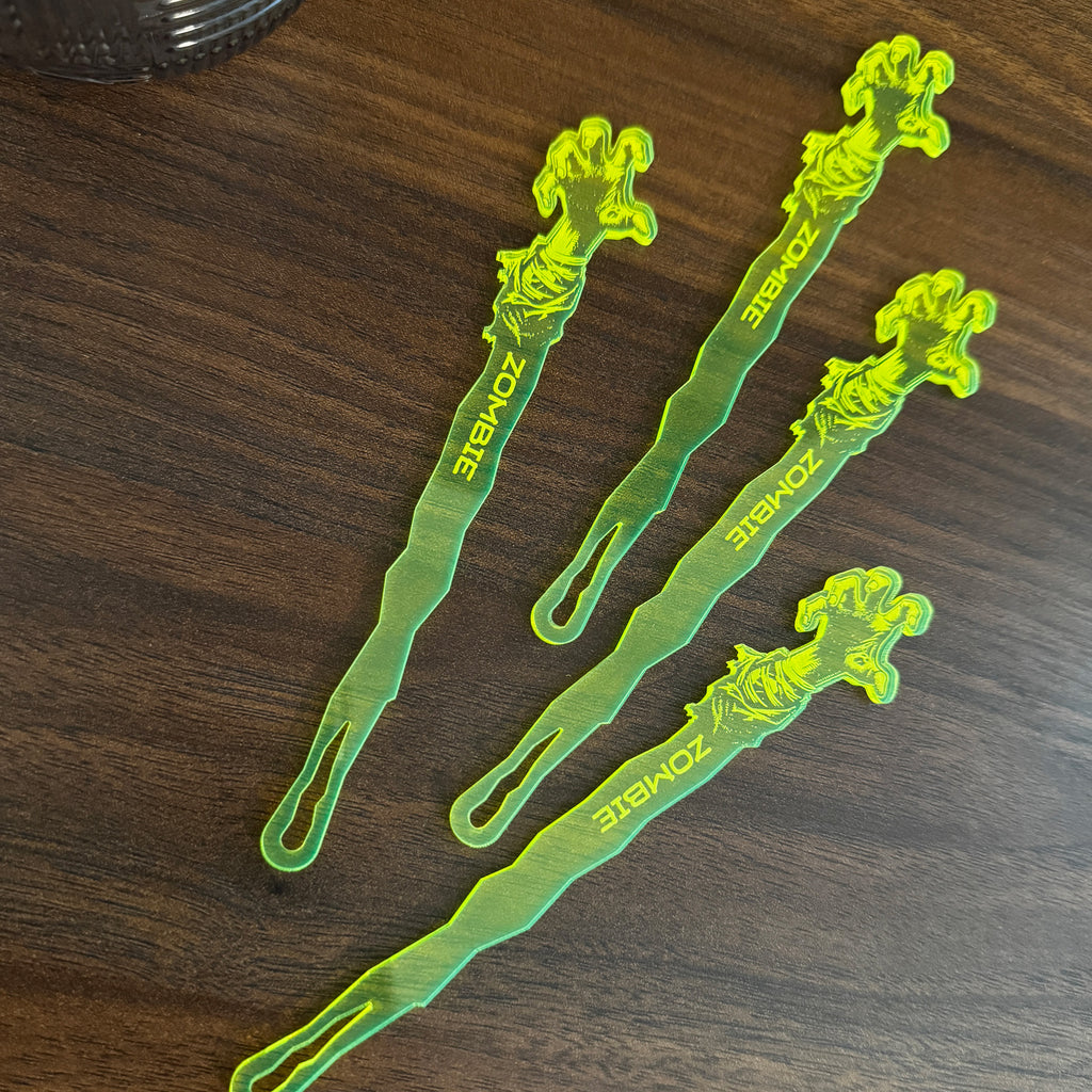 Zombie Personalized Stubbie Swizzle Sticks resting on a dark wooden table next to an glass tumbler.